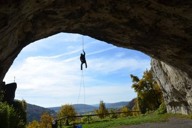 Eine Person seilt sich an einem Seil von der Decke einer großen Felsenhöhle ab, im Hintergrund sind blauer Himmel, Bäume und eine hügelige Landschaft zu sehen.