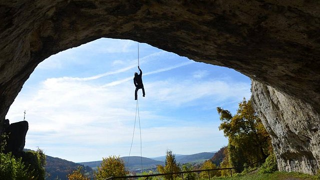Eine Person seilt sich an einem Seil von der Decke einer großen Felsenhöhle ab, im Hintergrund sind blauer Himmel, Bäume und eine hügelige Landschaft zu sehen.